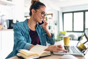Smiling business woman working with laptop while talking with smartphone in the kitchen at home.